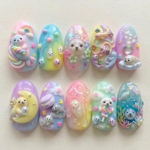 Op de afbeelding: Een set van tien kunstnagels, elk uniek ontworpen met pastelkleuren en 3D-versieringen. De ontwerpen tonen zeehonden, regenbogen, wolken en bloemen, wat een grillige en artistieke uitstraling cre&euml;ert. De nagels zijn ovaal van vorm en tonen een verscheidenheid aan texturen en details.
