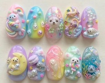 May include: A set of ten artificial nails, each uniquely designed with pastel colors and 3D embellishments. The designs feature seals, rainbows, clouds, and flowers, creating a whimsical and artistic aesthetic. The nails are oval-shaped and showcase a variety of textures and details.