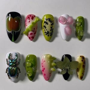 May include: A collection of ten uniquely designed artificial nails. The nails feature various designs, including fruit slices, insect embellishments, and abstract art. Colors include black, white, green, pink, and yellow, with intricate details and textures.