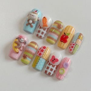 May include: A set of ten artificial nails with various designs. The nails feature cartoon monkeys, fish, apples, and patchwork patterns. Colors include pink, blue, yellow, and brown. Some nails have 3D embellishments like buttons and a butterfly.