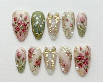 May include: A set of ten artificial nails with various designs. The nails feature hand-painted floral patterns, including roses and lily of the valley, with gold accents and pearl embellishments. Some nails have gold bow designs.