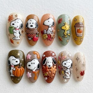 May include: A set of ten press-on nails featuring Snoopy and Woodstock in autumn-themed designs. The nails display various fall colors, glitter accents, pumpkins, leaves, and a coffee cup, with raised 3D details and star motifs on a glossy finish.