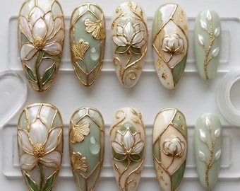 Könnte beinhalten: Ein Set aus zehn künstlichen Fingernägeln mit botanischem Motiv in Salbeigrün, Creme und irisierendem Weiß. Die Designs zeigen erhabene, gold umrandete Blumenmuster, Ginkgoblätter und zarte Ranken mit kleinen weißen Akzenten.