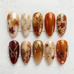 May include: A set of ten almond-shaped press-on nails featuring autumn-themed designs. The collection includes cream, orange, and deep burgundy bases decorated with gold leaf flakes, painted maple leaves, delicate branches, mushrooms, and small pearls.