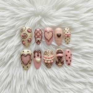 May include: A set of ten artificial nails with various designs. The nails feature a palette of pink, brown, and cream colors. Designs include hearts, bows, polka dots, and swirls, with some nails resembling ice cream cones.