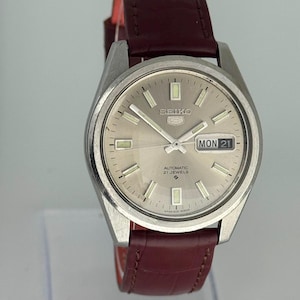 Puede incluir: Un reloj de pulsera automático Seiko 5 con esfera plateada efecto rayos de sol, índices de bastón plateados y ventana de día y fecha en la posición de las tres. El reloj cuenta con caja de acero inoxidable y correa de cuero burdeos oscuro.