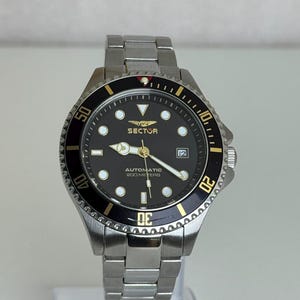 Può includere: Un orologio da polso automatico Sector in acciaio inossidabile con quadrante nero, lancette e indici color oro e una ghiera nera girevole con numeri color oro. Il quadrante riporta la scritta Sector, Automatic e 200 Meters con data.