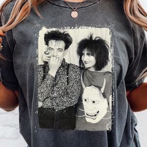 May include: Dark gray t-shirt featuring a black and white image of two people. The image is in a rectangular frame. The person on the left is wearing a patterned shirt and suspenders. The person on the right is wearing a red top and a white mask.