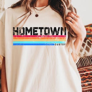 Puede incluir: Camiseta color crema con la palabra "HOMETOWN" en letras negras sobre un diseño de rayas arcoíris. La palabra "CUSTOM" está impresa debajo. La camiseta tiene un corte informal y de gran tamaño.