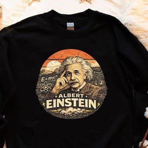 Puede incluir: Sudadera negra con un gráfico circular de Albert Einstein. El gráfico incluye la ecuación E=mc² y el texto "ALBERT EINSTEIN" sobre un fondo de puesta de sol. Diseño de estilo vintage.