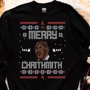 May include: Black sweatshirt with a festive design. The design includes the text "MERRY CHRITHMITH" in white, a portrait of Mike Tyson, and snowflake and reindeer patterns in red and white. A fun holiday sweater.