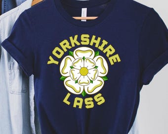May include: A navy blue t-shirt featuring the words "YORKSHIRE LASS" in yellow, encircling a white rose emblem with yellow and green details. The shirt is displayed on a wooden hanger with other garments.