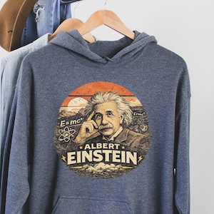 Puede incluir: Una sudadera con capucha azul marino con un gráfico circular de Albert Einstein. El gráfico incluye la ecuación E=mc² y el texto "ALBERT EINSTEIN". La sudadera con capucha está colgada en una percha de madera.