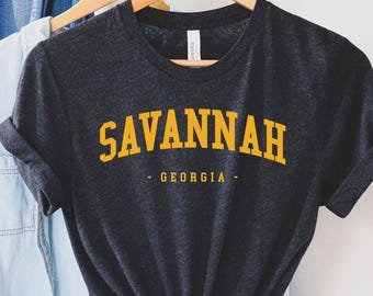 Puede incluir: Una camiseta gris oscuro con la palabra "SAVANNAH" en letras amarillas y "GEORGIA" debajo. La camiseta está colgada en una percha de madera con otras prendas, incluyendo una camisa vaquera y un sombrero marrón.