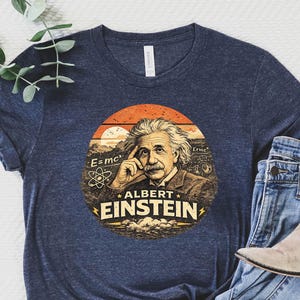 May include: Navy blue t-shirt featuring a vintage-style graphic of Albert Einstein with the text "E=mc²" and "Albert Einstein". The shirt is paired with blue jeans and beige ankle boots, creating a casual, everyday look.