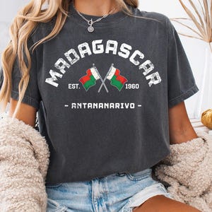 Può includere: T-shirt grigio scuro con la scritta "MADAGASCAR" ad arco, con il testo "EST. 1960 - ANTANANARIVO" sotto. Due bandiere incrociate sono al centro. La maglietta è indossata con pantaloncini di jeans.