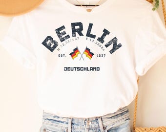 Puede incluir: Una camiseta blanca con la palabra "BERLIN" en azul marino, la bandera alemana y la palabra "DEUTSCHLAND". El conjunto incluye unos shorts vaqueros desgastados, un sombrero beige y un bolso bandolera tejido.