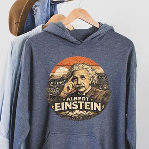 Puede incluir: Una sudadera con capucha azul marino con un gráfico circular de Albert Einstein. El gráfico incluye el texto "ALBERT EINSTEIN", la fórmula E=mc² y un símbolo de átomo. La sudadera con capucha está colgada en una percha de madera.