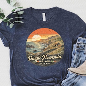 May include: Navy blue t-shirt featuring a scenic sunset design over the Dingle Peninsula in Ireland. The graphic includes a coastal landscape with a cottage. The shirt is paired with blue jeans and beige ankle boots.