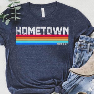 以下が含まれることがあります： 「HOMETOWN」の白ブロック文字と水平レインボーデザインが特徴のネイビーブルーTシャツ。「CUSTOM」の文字が下にあります。青いジーンズとタンカラーのアンクルブーツと組み合わせています。