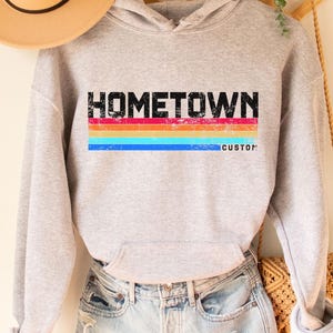 Könnte beinhalten: Ein hellgrauer Hoodie mit dem Wort "HOMETOWN" in schwarzen Blockbuchstaben über horizontalen Streifen in Rot, Orange, Gelb, Blau und einem benutzerdefinierten Wort in Blau. Ein beigefarbener Hut und Jeansshorts sind ebenfalls im Bild.