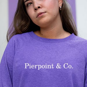 Op de afbeelding: Een paarse sweatshirt met de witte tekst "Pierpoint & Co.". De sweatshirt is effen van kleur en heeft een ronde hals. De persoon op de afbeelding draagt de sweatshirt.