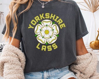 May include: Charcoal grey t-shirt with a white rose emblem and the words "Yorkshire Lass" in yellow. The casual tee is suitable for everyday wear. The design is centered on the front of the shirt, a popular style in the UK.