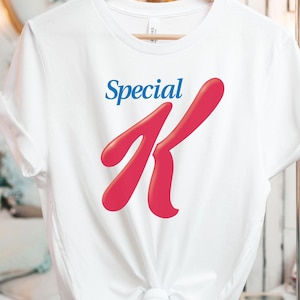 Puede incluir: Una camiseta blanca con las palabras "Special K" en azul y rojo. La "K" roja es estilizada y prominente. La camiseta está colgada en una percha de madera.