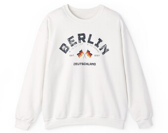 Puede incluir: Sudadera blanca con la palabra "BERLIN" arqueada en el pecho en una fuente gris oscuro desgastada. Debajo, dos pequeñas banderas y la palabra "DEUTSCHLAND". La sudadera tiene cuello redondo y mangas largas.