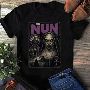 Puede incluir: Una camiseta negra con un estampado gráfico del póster de la película La Monja. El diseño incluye un retrato central de la monja, dos viñetas circulares que muestran un edificio gótico y un pasillo oscuro, y el texto The Nun y Witness the darkest chapter in The Conjuring.