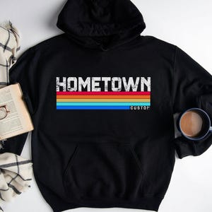 May include: Black hoodie featuring the word "HOMETOWN" in a distressed white font above a horizontal stripe design in orange, yellow, red, and blue. The word "CUSTOM" is below the stripes. A book, glasses, and a plaid scarf are in the background.