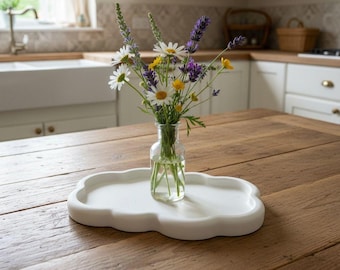 May include: A small glass vase filled with wildflowers, including daisies and lavender, sits on a white cloud-shaped tray. The tray rests on a wooden table. The kitchen in the background has white cabinets and a farmhouse sink.
