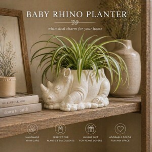 May include: A cream-colored rhino-shaped planter holding a green and white plant, resting on a wooden shelf. The planter has a detailed design and is labeled "BABY RHINO PLANTER". The shelf also holds books and a vase.