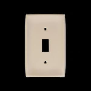 May include: Cream-coloured light switch cover with a rectangular switch opening and two circular screw holes. The cover has rounded corners and a smooth finish, set against a black backdrop.