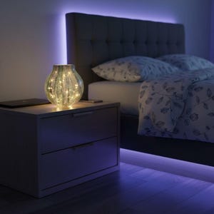 May include: A decorative glass vase with internal fairy lights sits on a nightstand next to a bed. The vase has a textured, green and silver design. The bed has a gray headboard and floral patterned bedding. The room is illuminated with blue LED lighting.