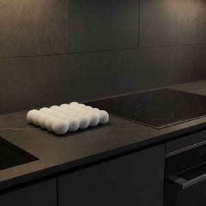Peut inclure: Un porte-œufs blanc, avec des alvéoles sphériques, est posé sur un plan de travail sombre, à côté d'une plaque de cuisson noire. La cuisine présente des meubles sombres et une crédence assortie, créant une esthétique moderne et minimaliste.