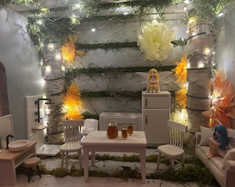 May include: A miniature fairy-themed dollhouse interior with a kitchen, living room, and bathroom. The walls are decorated with birch bark, moss, and fairy lights. A small table is set with drinks. A fairy figurine sits on a small refrigerator.