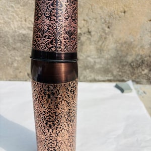 May include: A copper-coloured water bottle featuring an elaborate, swirling floral pattern. The bottle has a dark brown cap and base, with the design etched into the metal. The cylindrical bottle appears to be made of metal.