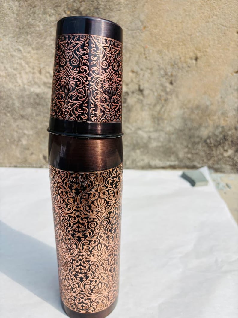 May include: A copper-coloured water bottle featuring an elaborate, swirling floral pattern. The bottle has a dark brown cap and base, with the design etched into the metal. The cylindrical bottle appears to be made of metal.