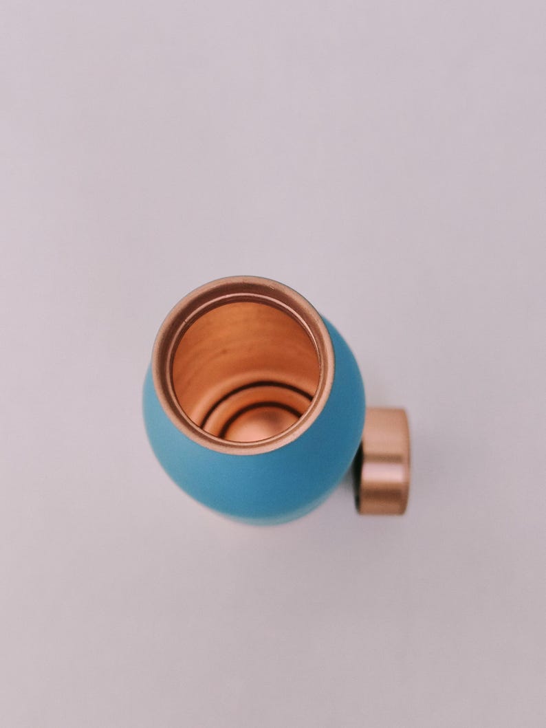 May include: A teal water bottle with a copper interior and a copper cap. The bottle has a rounded shape and a smooth finish. The cylindrical cap is attached to the side. The bottle is designed for carrying liquids.