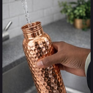 May include: A cylindrical copper water bottle with a textured, hammered finish. The bottle has a narrow neck and a metallic rim, and it is being filled with water from a stainless steel kitchen faucet, showing the reflective surface of the metal.