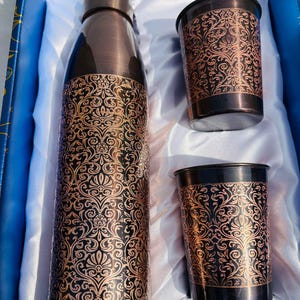 May include: A set of copper-coloured water bottle and two tumblers, featuring an intricate floral pattern. The items are presented in a white-lined box. The bottle and tumblers have a dark brown base and rim, with a lighter copper-toned, detailed design.