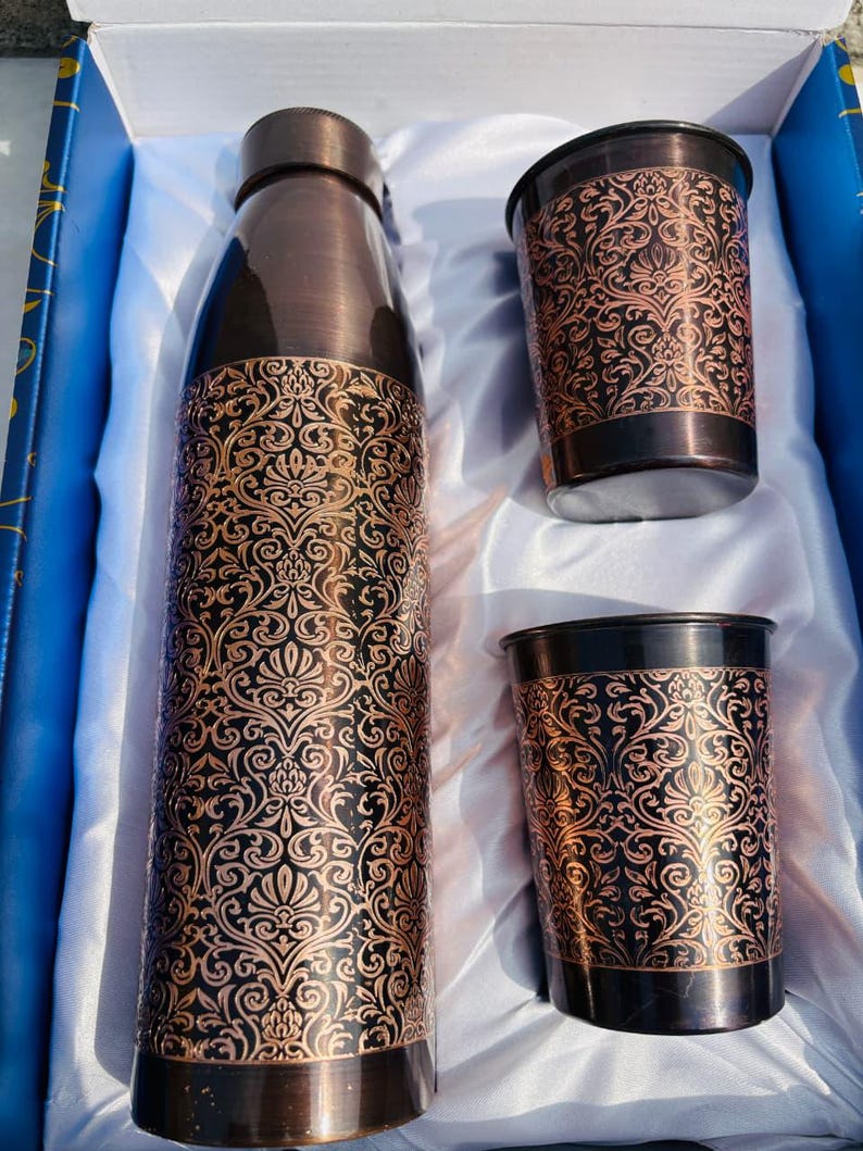 May include: A set of copper-coloured water bottle and two tumblers, featuring an intricate floral pattern. The items are presented in a white-lined box. The bottle and tumblers have a dark brown base and rim, with a lighter copper-toned, detailed design.