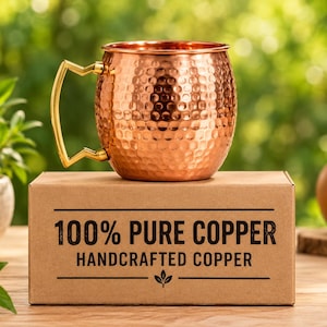 May include: A hammered copper mug with a gold-toned angular handle sits atop a rectangular cardboard box. The box features black text that reads 100% PURE COPPER HANDCRAFTED COPPER with a small leaf icon centred below the text.