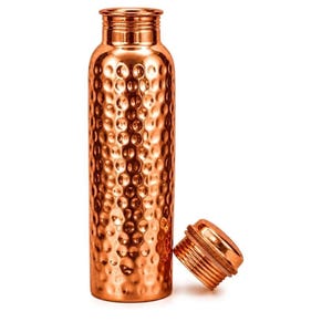 May include: A cylindrical copper water bottle with a polished, hammered texture across its surface. The bottle features a narrow neck and a matching screw-top lid, which is shown detached and resting beside the base of the bottle.