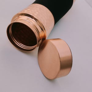 May include: A copper water bottle with a hammered texture and a matching lid. The bottle has a black sleeve and is partially open, revealing the interior. The lid is placed next to the bottle.