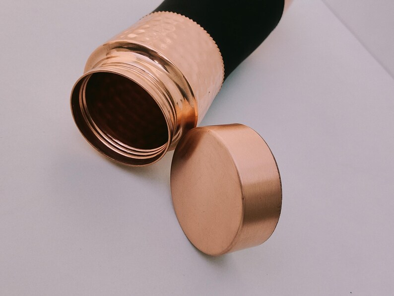 May include: A copper water bottle with a hammered texture and a matching lid. The bottle has a black sleeve and is partially open, revealing the interior. The lid is placed next to the bottle.
