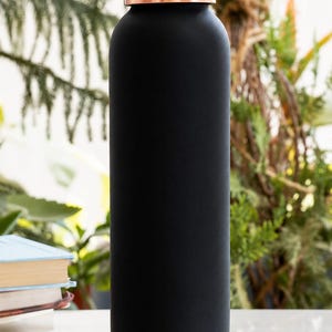 May include: A cylindrical water bottle with a matte black finish, featuring a metallic copper-coloured screw-top lid and a matching copper-coloured base. The bottle has a smooth, uniform texture and a straight-sided design with a rounded top.