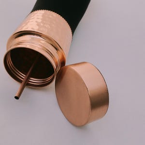 May include: A copper-colored water bottle with a black top and a matching lid. The bottle has a hammered texture on the copper section and a straw inside. The lid is off to the side.