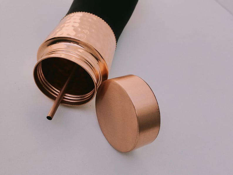May include: A copper-colored water bottle with a black top and a matching lid. The bottle has a hammered texture on the copper section and a straw inside. The lid is off to the side.